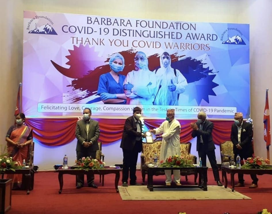 Dr. Niraj Kumar Singh receiving the Barbara Foundation COVID-19 Distinguished Award on stage beneath a banner reading “Thank You COVID Warriors”.